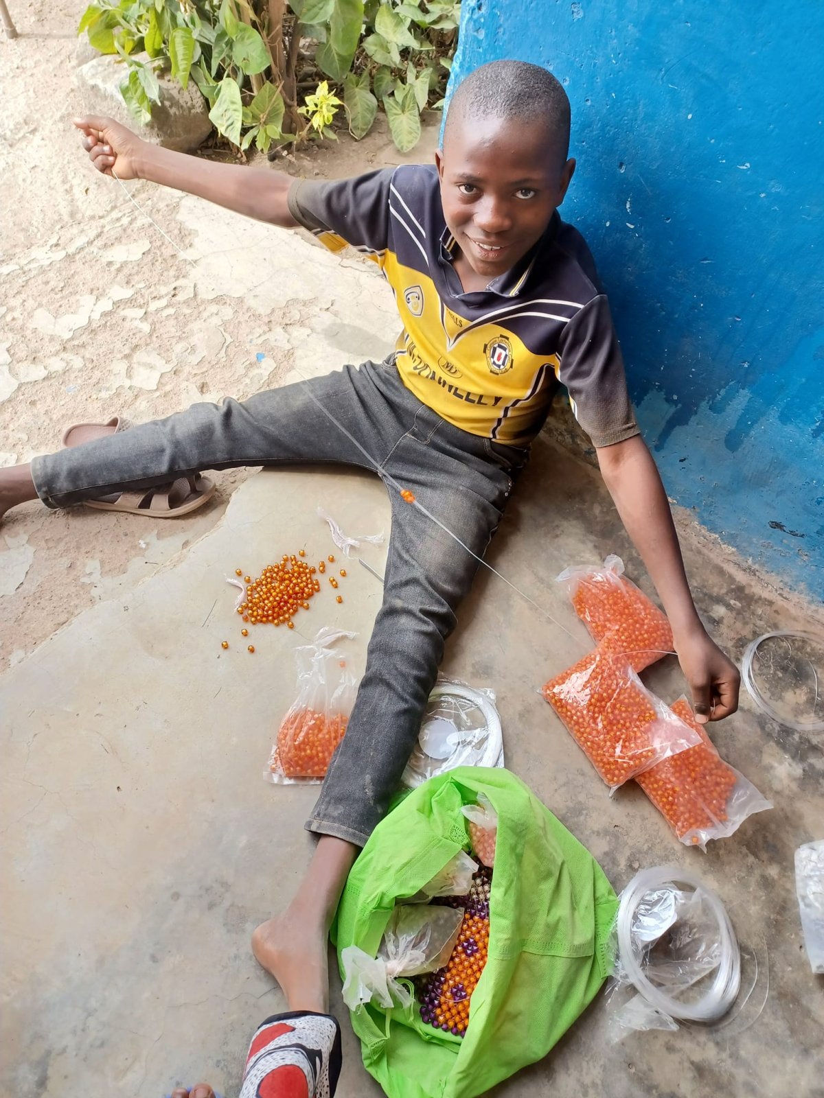 A warmly smiling boy sitting on the floor, happily focused on threading beads to create his artwork, hoping to shape his skills and showcase his talent for a brighter future.